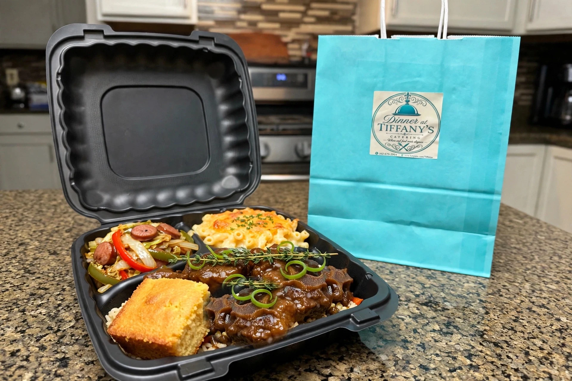 Braised oxtails over rice with a square of cornbread, mac and cheese and fried cabbage in a black to-go container on a kitchen counter, the Tiffany blue Dinner at Tiffany's bag standing beside it